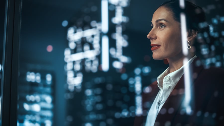 A woman standing in an office window, with city lights reflecting