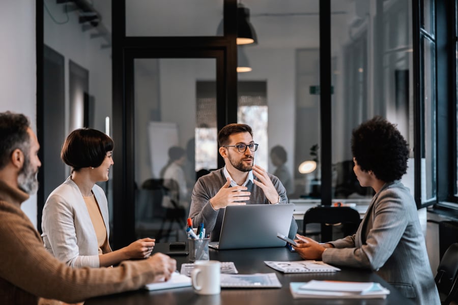 A meeting room in an office where four coworkers sit around a table and listen to a man speak in front of an open laptop