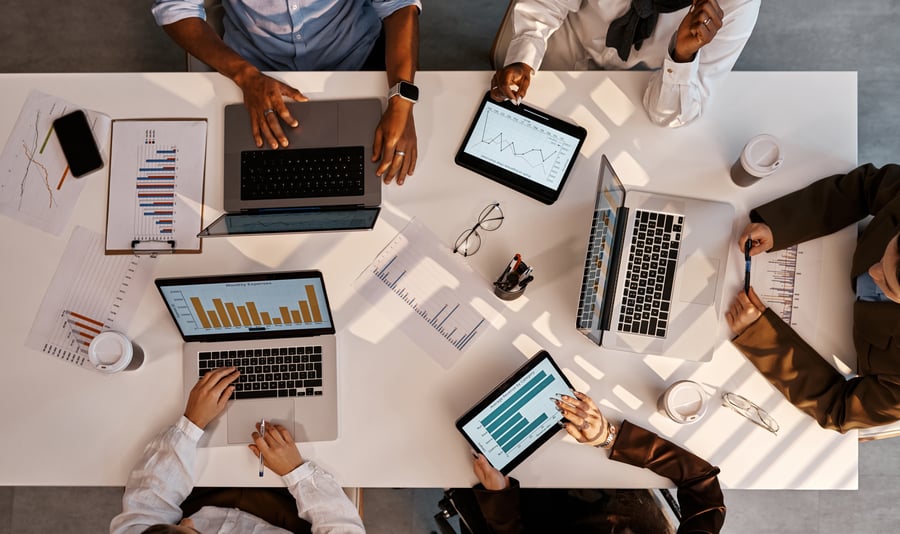 An overhead shot of a table in an office, with four coworkers sitting around a white table with laptops and tablets open to graphs and charts