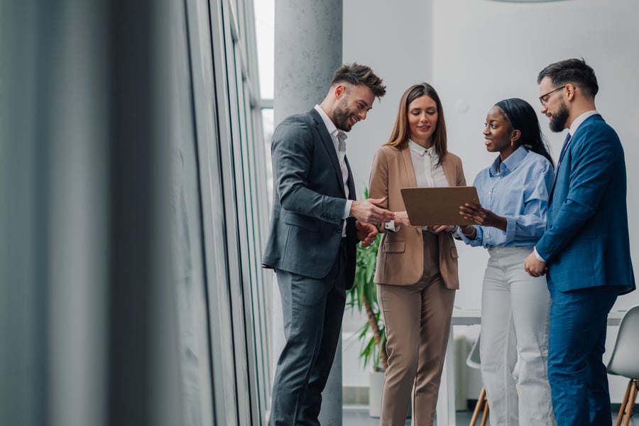 A group of four coworkers standing in a brightly-lit office, smiling around a tablet