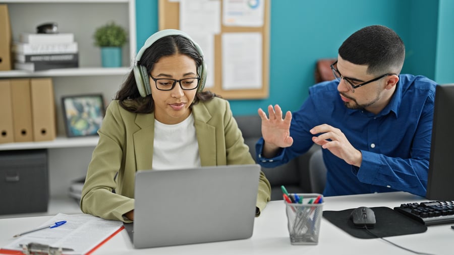 A woman typing on a laptop wearing headphones while a man tries to speak to her in an office