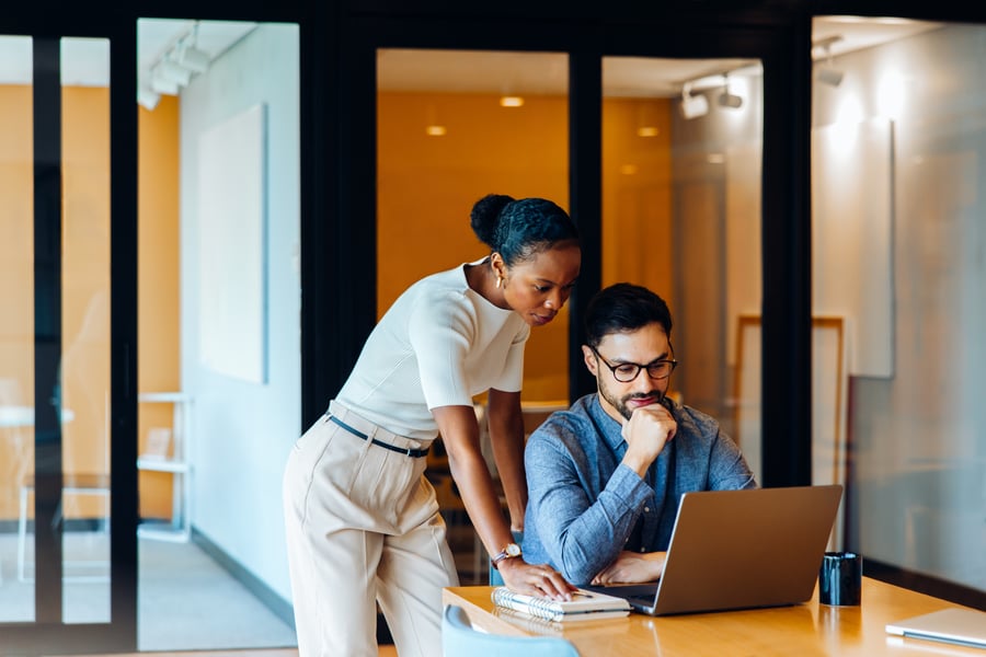 A woman standing over a man's shoulder to read something on his laptop in an office