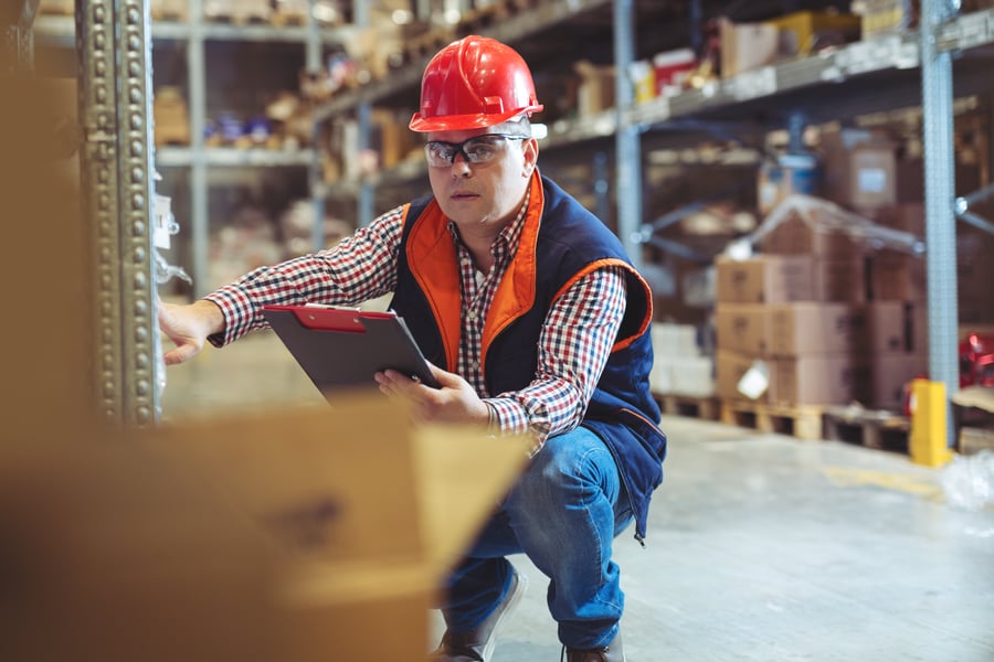 A man squats in a warehouse, checking something on a clipboard. He's wearing a red hard hat and orange vest.