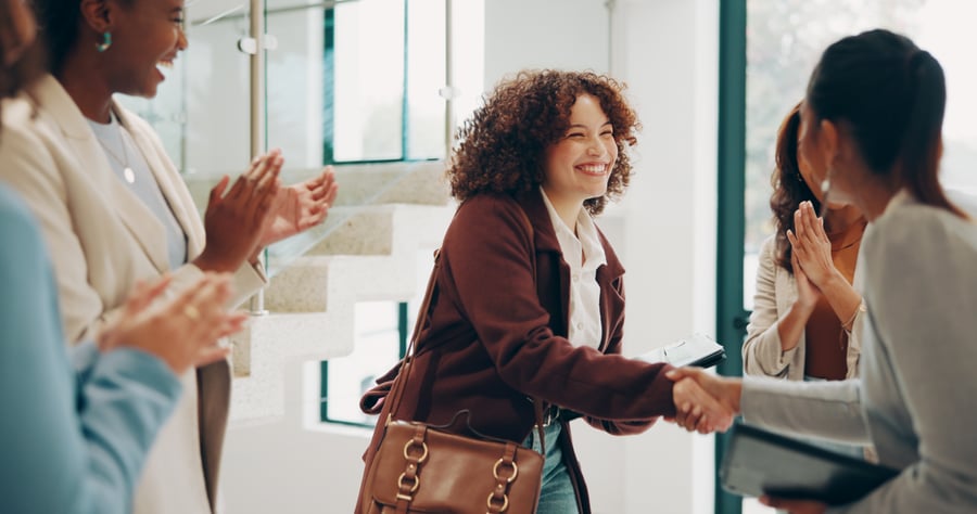 A woman in a business setting smiling and shaking hands with someone as the people around them clap