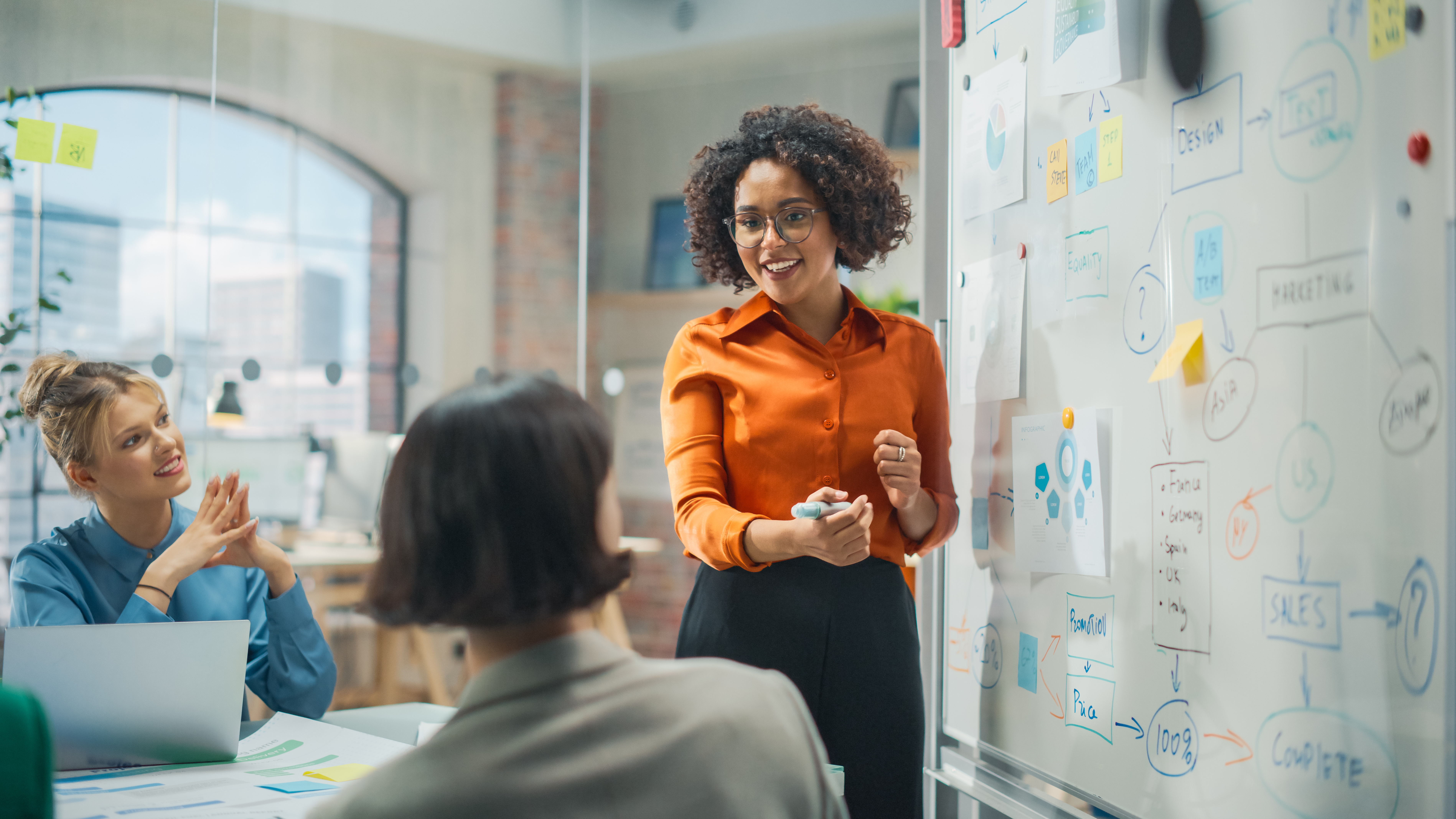 A woman standing by a whiteboard in an office, going over notes